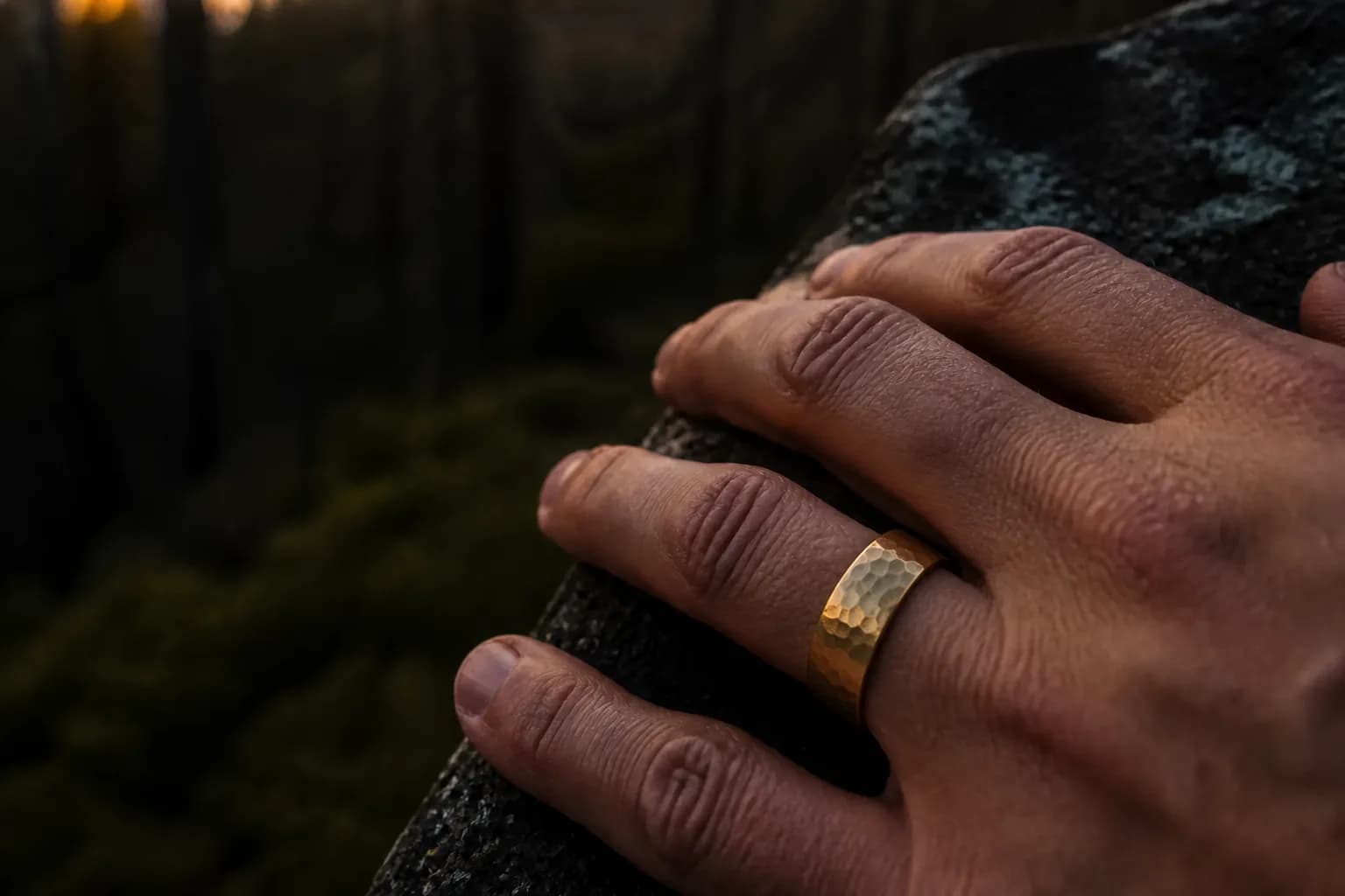 A man's hand resting on weathered granite at dusk in a pine forest, wearing a wide hammered gold tungsten wedding band on his ring finger