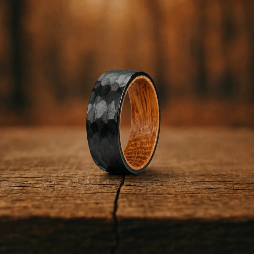 The Ember, an 8mm hammered black tungsten carbide band with a whiskey barrel wood inlay, standing upright on a weathered oak plank