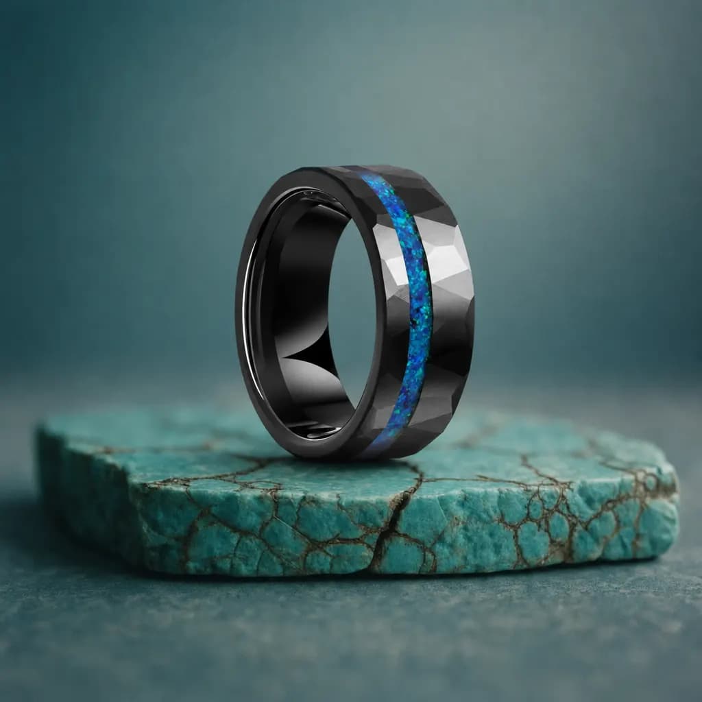 The Spectra, an 8mm faceted tungsten carbide band in black and blue with an opal inlay, standing upright on a turquoise veined mineral slab