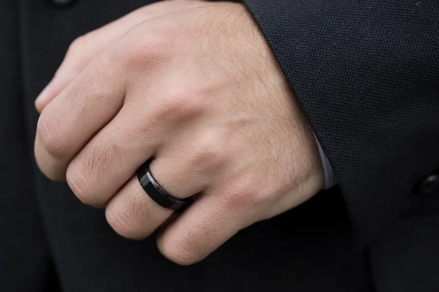 A black tungsten band worn on a hand against a dark suit cuff